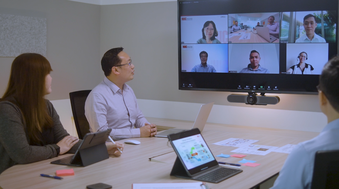 Three people are seated at a conference table participating in a video call displayed on a wall-mounted screen.